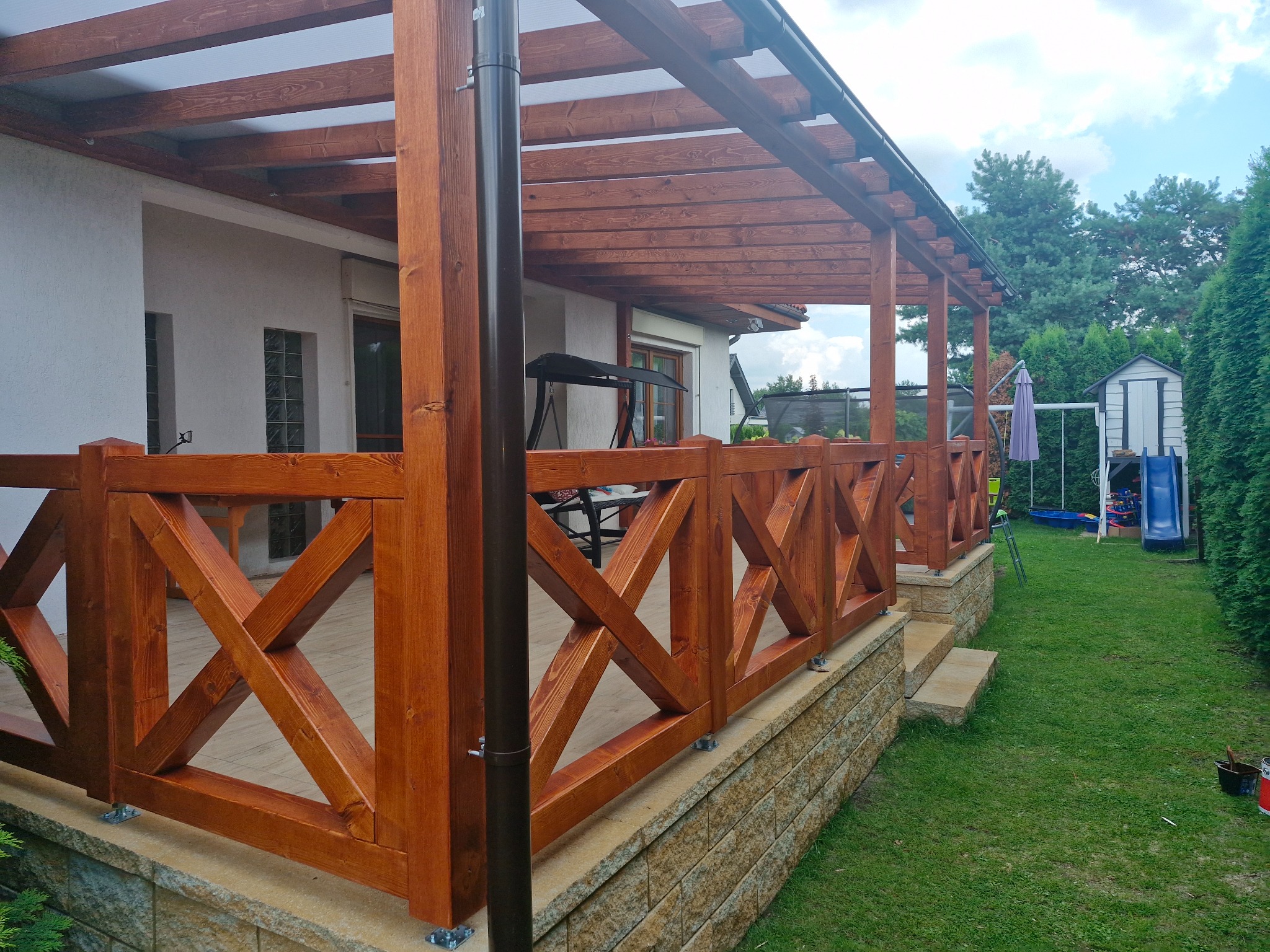 Drewniana pergola z balustradą o krzyżowym wzorze, osłaniająca taras. W tle widoczne drzewa i elementy ogrodu. Jasne drewno, solidna konstrukcja.