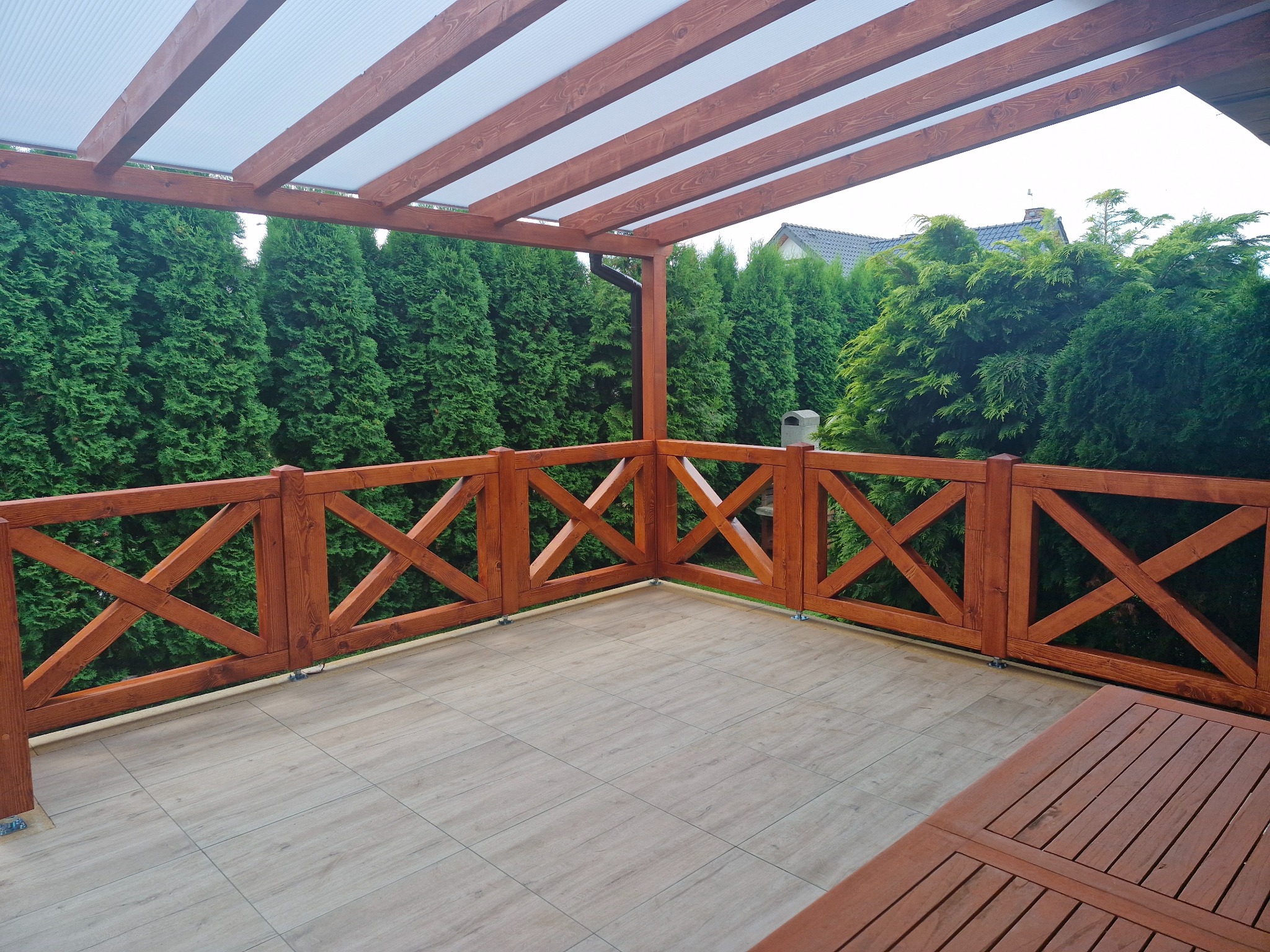 Zadaszony taras z drewnianą pergolą i balustradą krzyżową, wyłożony płytkami imitującymi drewno. W tle wysokie tuje.