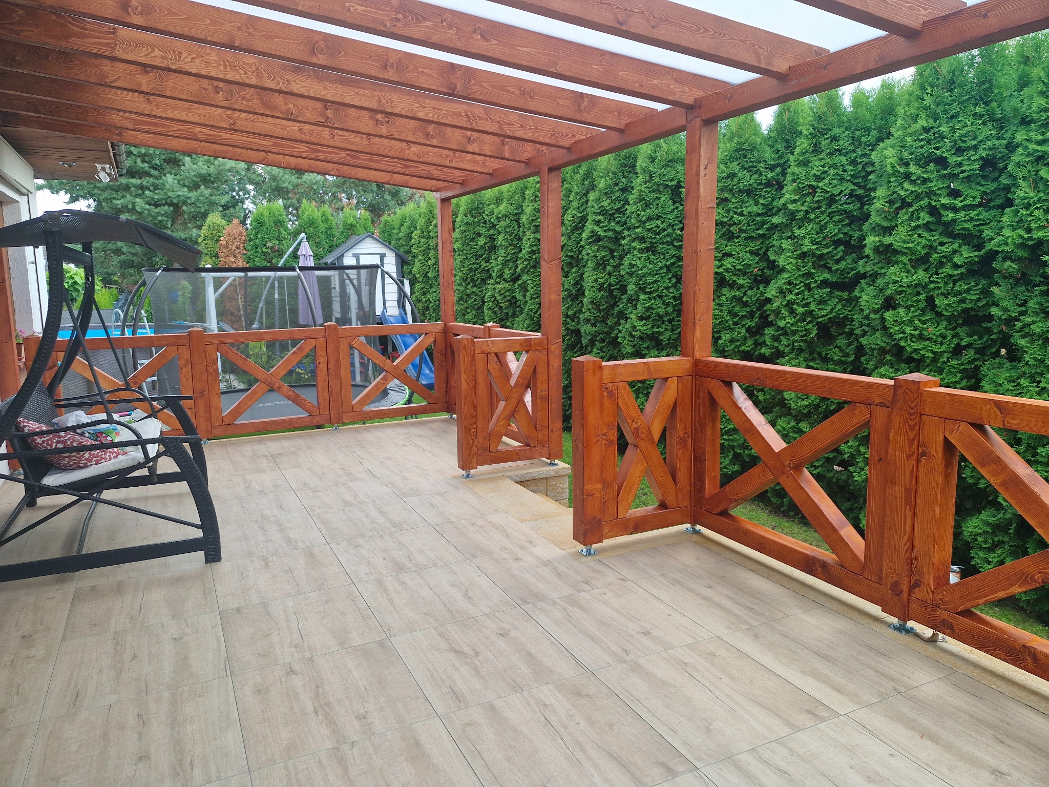 Zadaszony taras z drewnianą pergolą i balustradą, wykończony jasnymi płytkami. W tle zielone tuje i huśtawka ogrodowa.