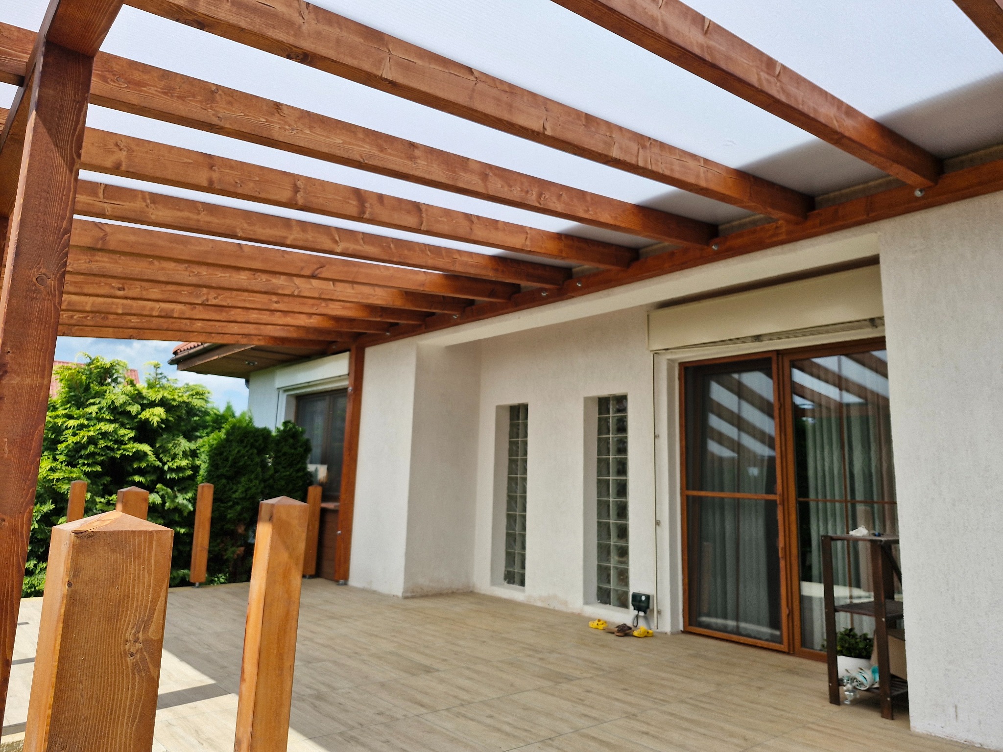 Drewniana pergola z jasnym zadaszeniem przylegająca do białego budynku. Widoczne elementy konstrukcyjne i fragment tarasu z płytkami.