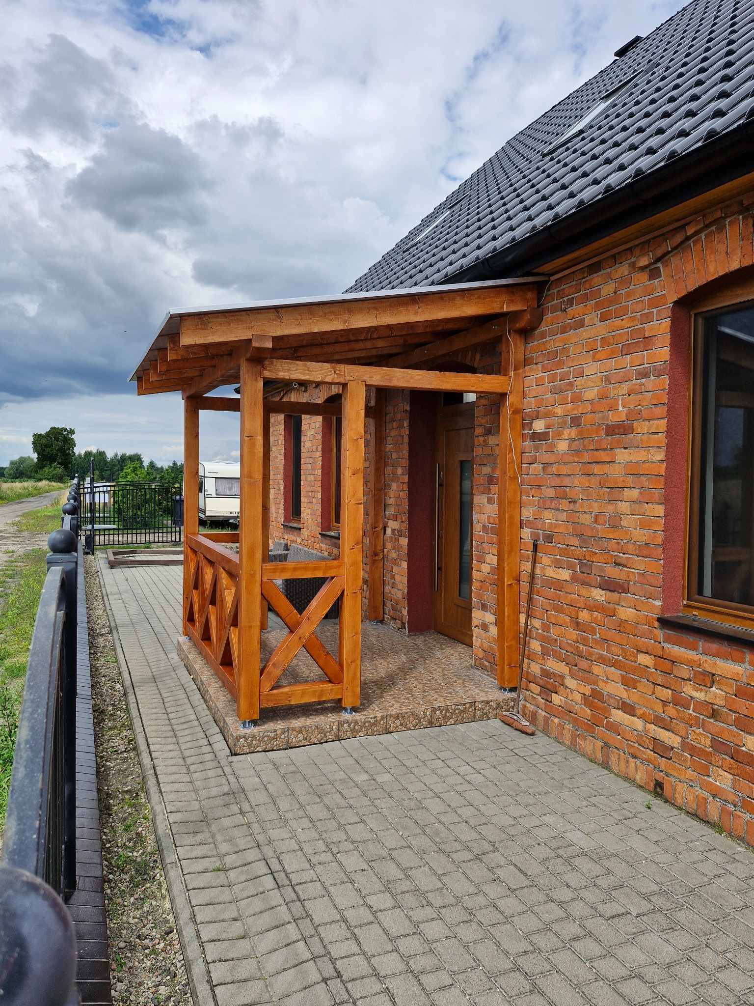 Drewniana pergola z balustradą przy ceglanym domu, widok z boku. Na pierwszym planie kostka brukowa i czarne metalowe ogrodzenie. W tle zieleń i przyczepa kempingowa.