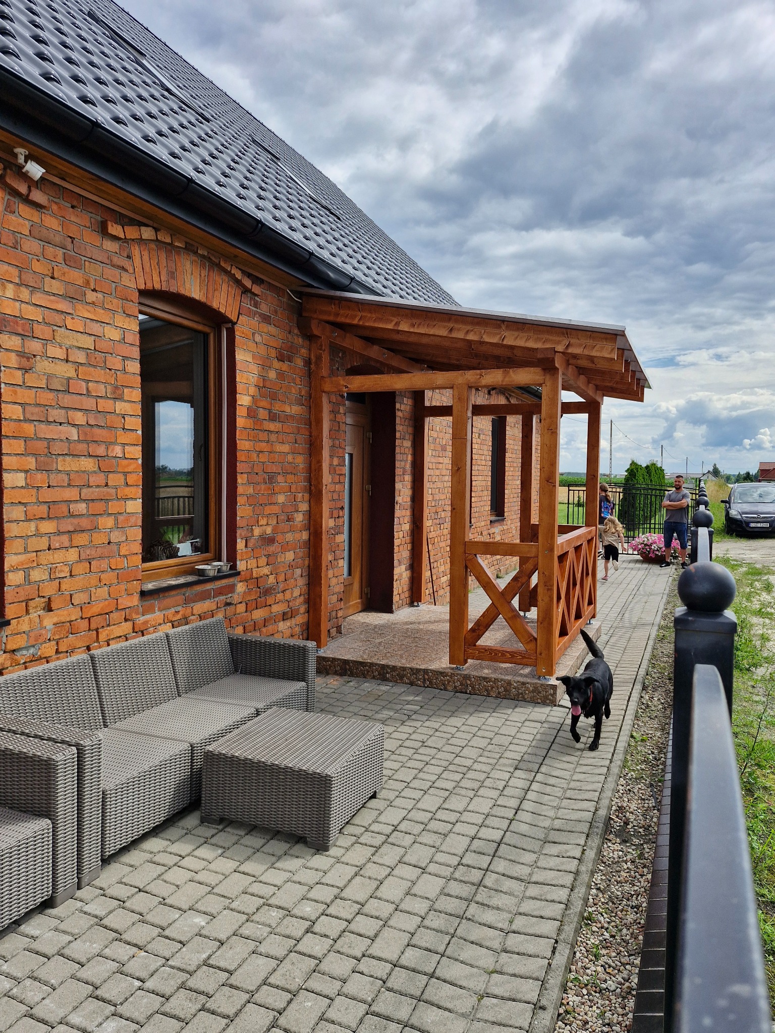 Ceglany dom z drewnianą pergolą i szarą sofą na kostce brukowej. W tle widoczni ludzie i samochód. Czarny pies biegnie w stronę widza.