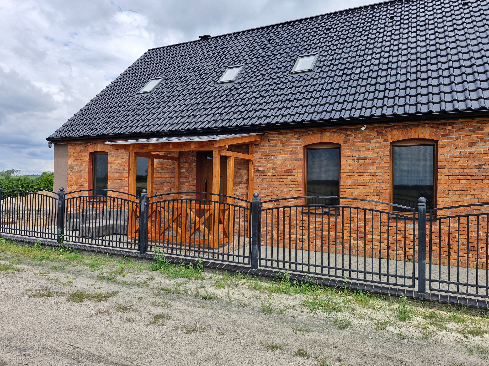 Dom z czerwonej cegły z czarną, metalową balustradą i drewnianą pergolą przy wejściu, w pochmurny dzień. Widoczne okna dachowe i fragment podjazdu.