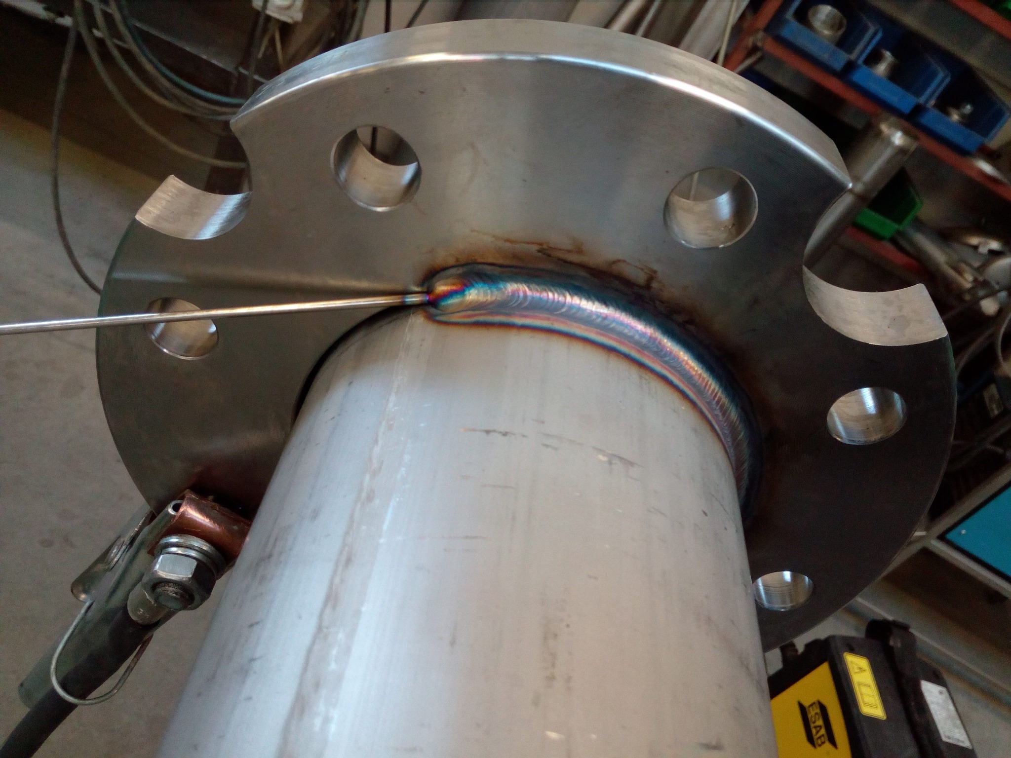 Zbliżenie na proces spawania kołnierza do rury ze stali nierdzewnej, widoczny łuk spawalniczy i tęczowe zabarwienie spoiny, uchwyt spawalniczy przymocowany do rury.