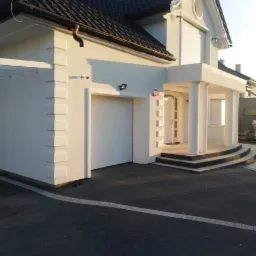 Biała elewacja domu jednorodzinnego z kolumnami przy wejściu i garażem, czarny dach, oświetlenie zewnętrzne.