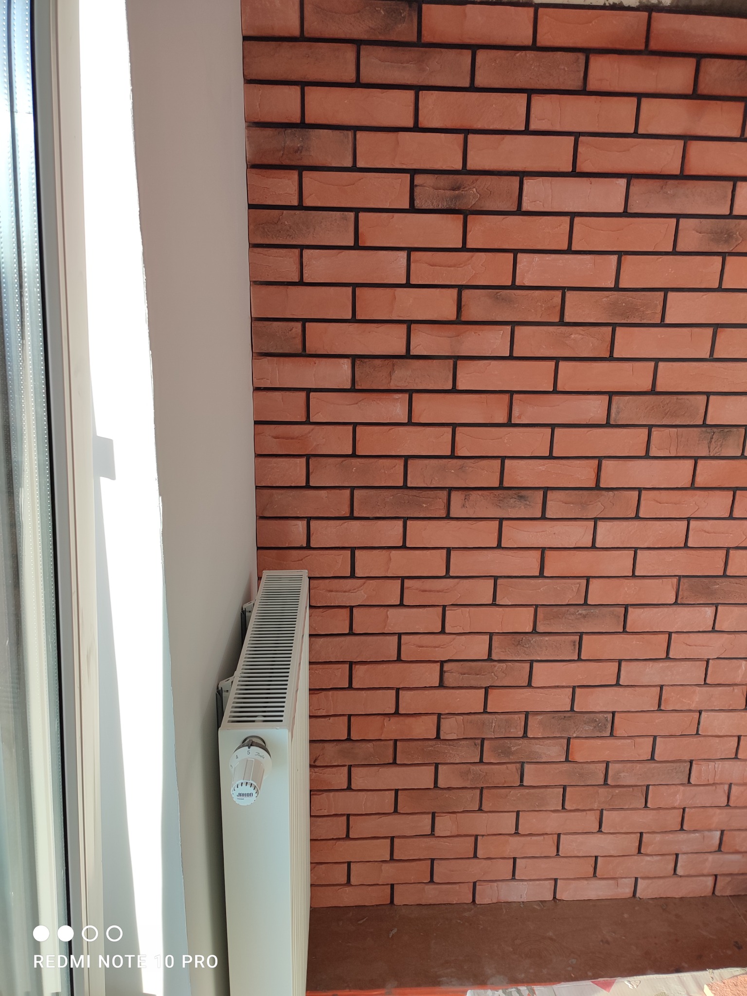 Ściana wykończona cegłą dekoracyjną w odcieniach czerwieni i brązu, sąsiadująca z białym grzejnikiem i oknem.