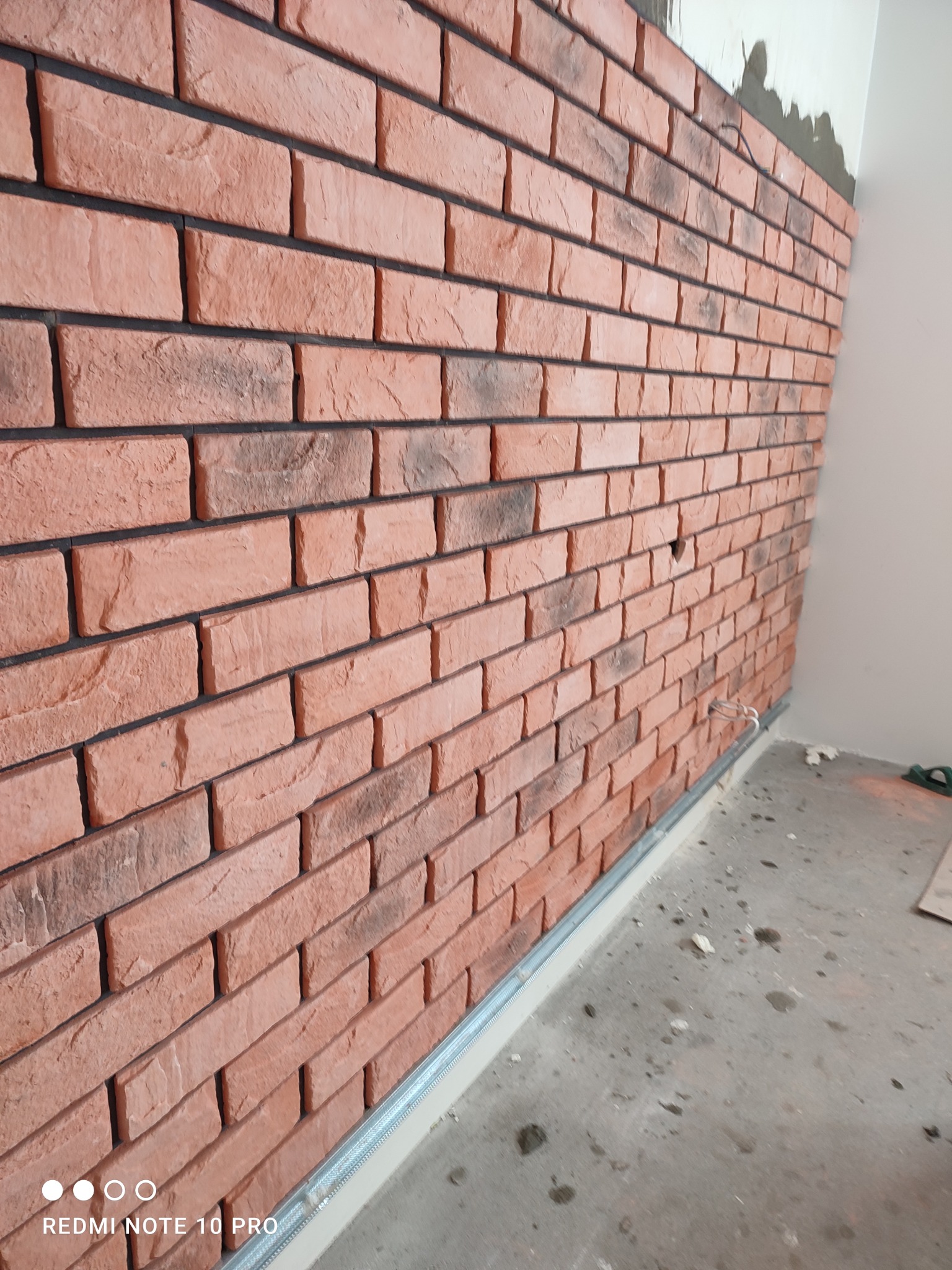 Ściana wyłożona dekoracyjną cegłą w odcieniach czerwieni i brązu, widoczna metalowa listwa przypodłogowa i fragment surowej ściany z resztkami zaprawy, podłoga z zabrudzeniami.