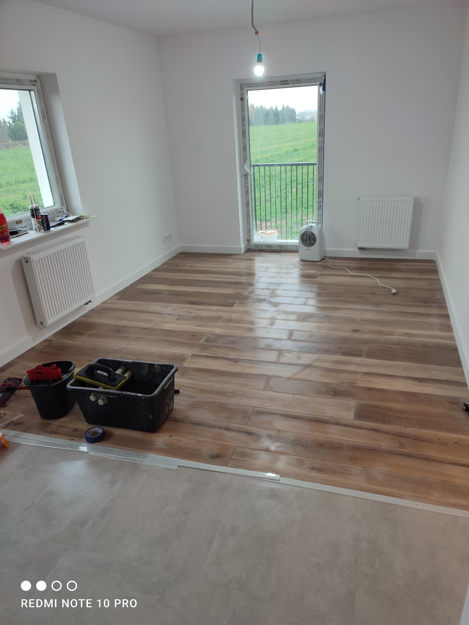 Wnętrze pokoju z ułożoną drewnopodobną podłogą; widoczne narzędzia glazurnicze, grzejnik i okno z widokiem na zieleń.