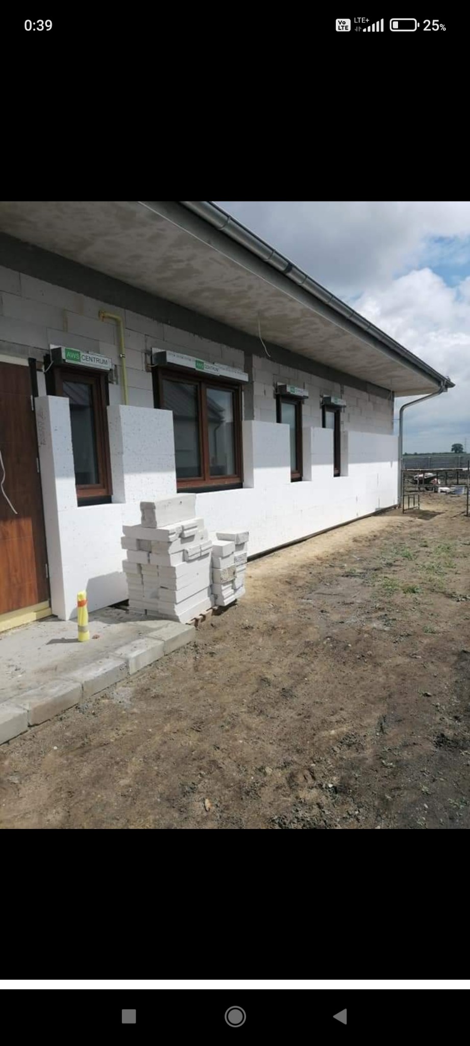 Ocieplanie ścian zewnętrznych styropianem w Szamotułach, widoczne okna z roletami i stos płyt styropianowych przy budynku.