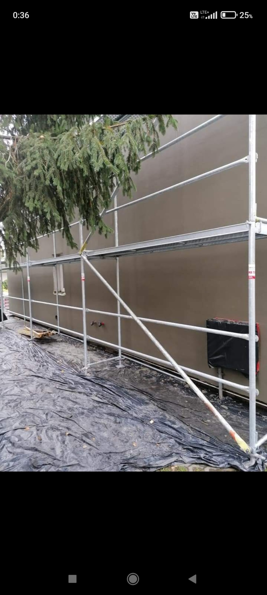 Elewacja budynku po nałożeniu gładzi, zabezpieczona folią i otoczona rusztowaniem; iglaste drzewo zwisa nad rusztowaniem.