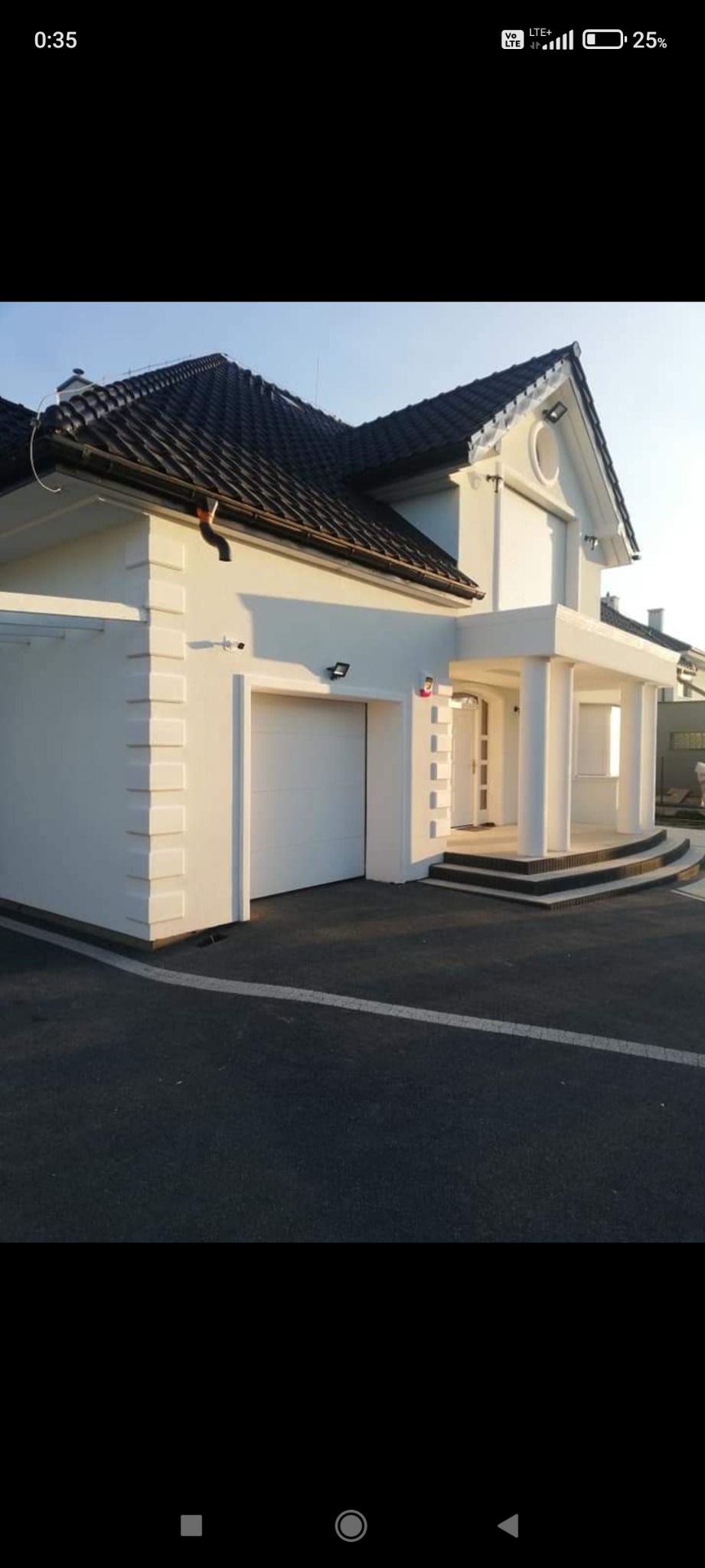 Biała elewacja domu jednorodzinnego z kolumnami przy wejściu i garażem, czarny dach, oświetlenie zewnętrzne.