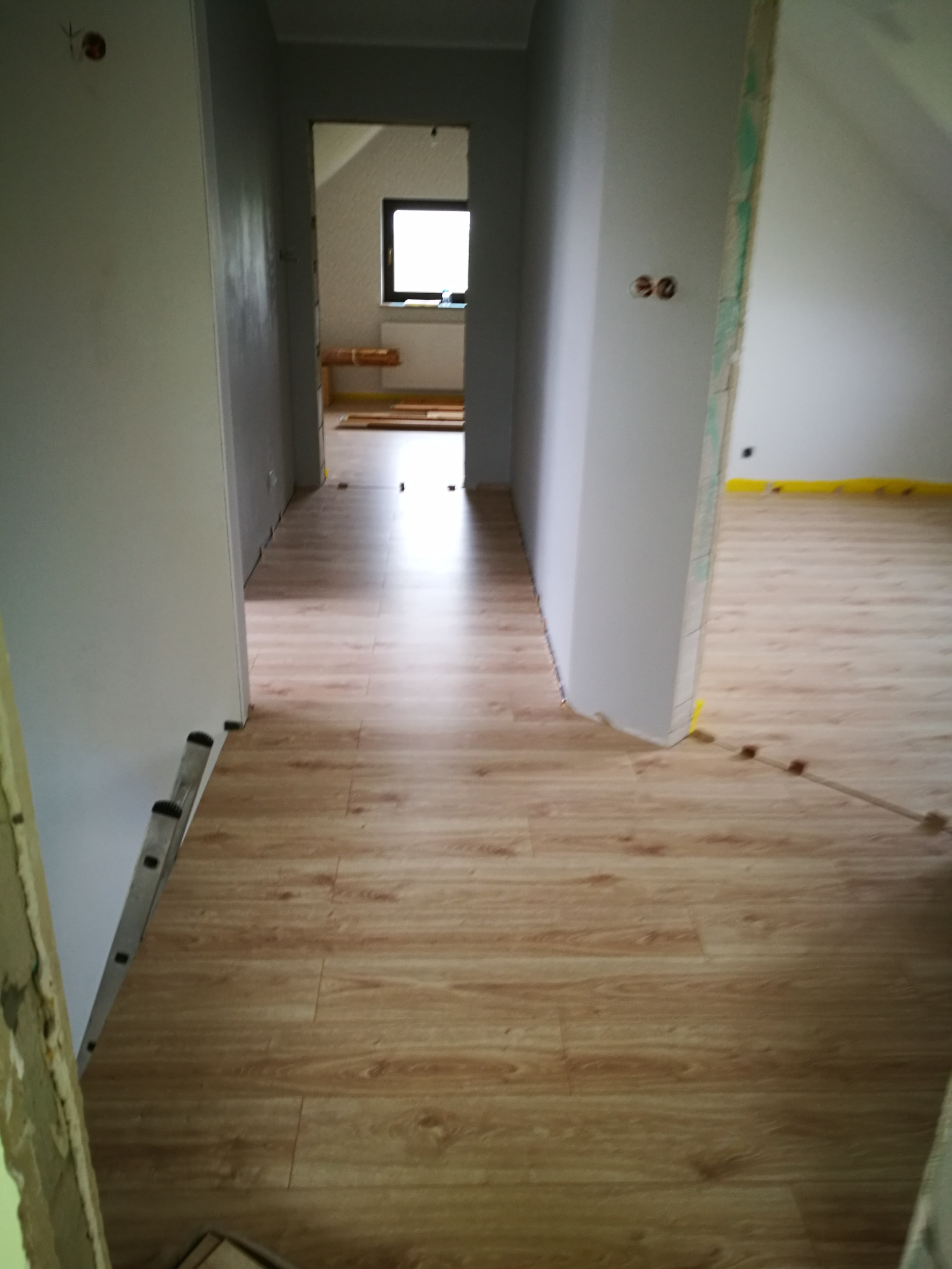 Nowa podłoga panelowa w trakcie układania, widoczne listwy przypodłogowe i drabina oparta o ścianę, przygotowania do dalszych prac wykończeniowych.