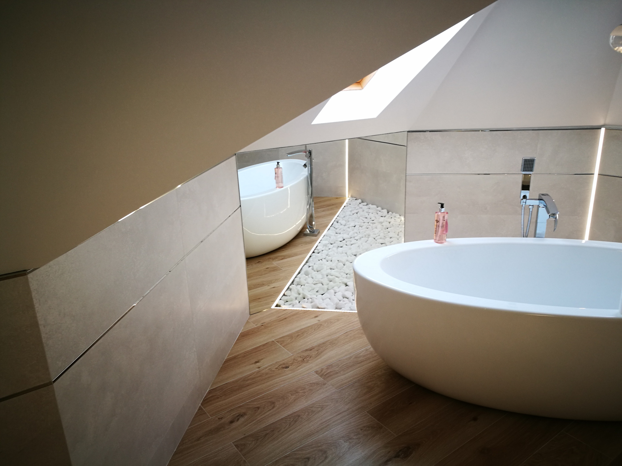 Nowoczesna łazienka na poddaszu z wolnostojącą wanną, drewnopodobną podłogą, białymi kamieniami dekoracyjnymi i lustrem odbijającym drugi plan.