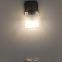 Świecąca, kwadratowa lampa ścienna z mlecznego szkła, zamontowana na drewnianej podstawie, widoczna nad gniazdkiem elektrycznym z włącznikiem, na gładkiej, białej ścianie.