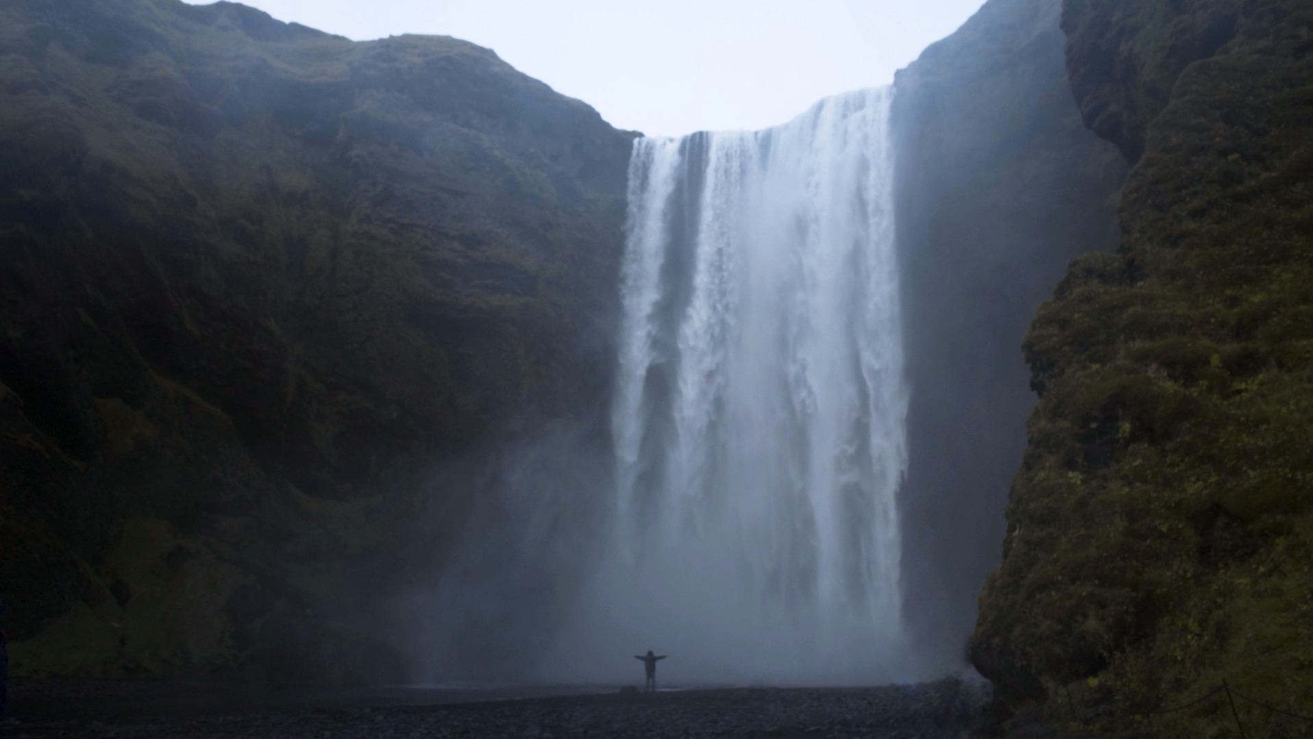 Szeroki kadr wodospadu Skógafoss w Islandii z osobą stojącą u jego podnóża z rozłożonymi ramionami, otoczonego skalistymi klifami porośniętymi mchem, w pochmurny dzień.