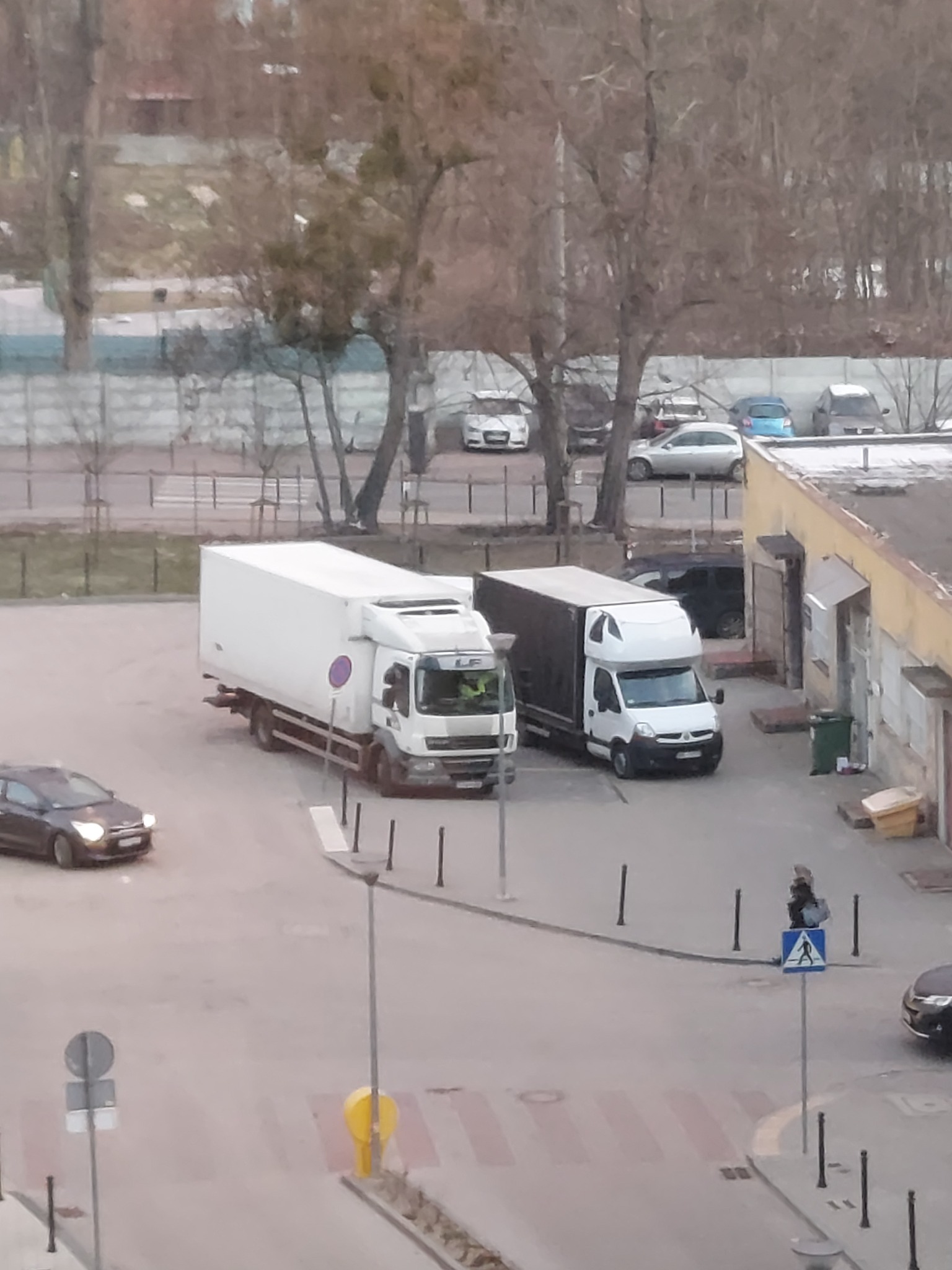Dwa busy transportowe zaparkowane na placu miejskim, jeden z białą skrzynią ładunkową, drugi z ciemną, widok z góry na otoczenie miejskie w pochmurny dzień.
