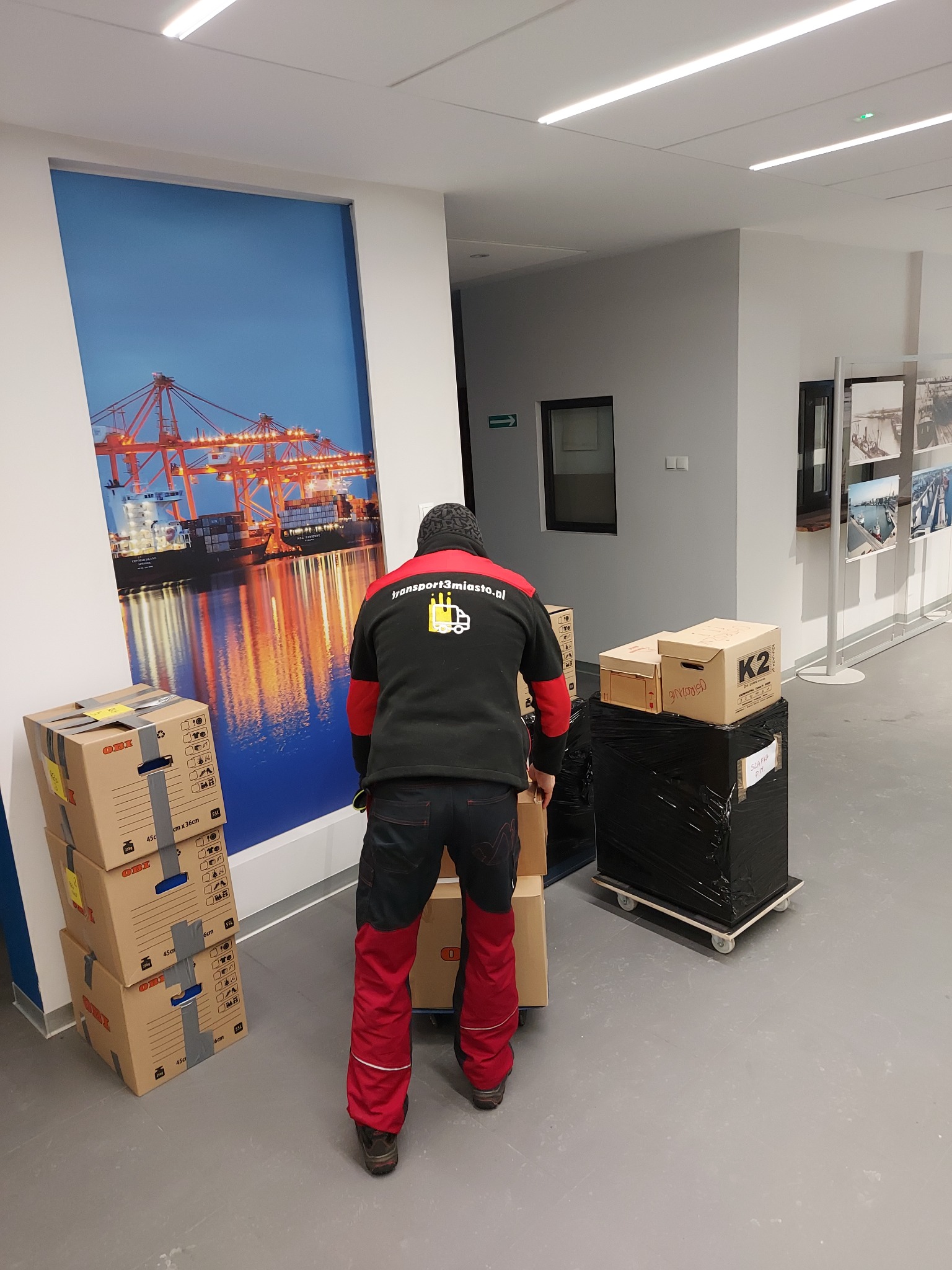 Pracownik w czerwono-czarnym stroju transportuje kartony w biurowym wnętrzu, w tle widoczna grafika z dźwigami portowymi.