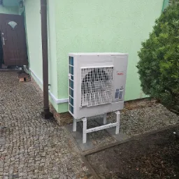 Zewnętrzna jednostka pompy ciepła marki Atlantic, zainstalowana na białym stelażu przy zielonej ścianie budynku w Białymstoku, widoczna kostka brukowa i iglak.