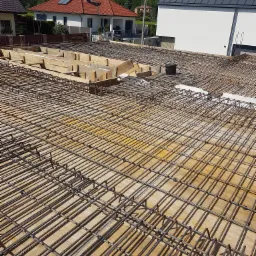 Zbrojenie stropu na deskowaniu, widok z góry na przygotowaną konstrukcję z prętów stalowych przed zalaniem betonem, w tle budynki mieszkalne.