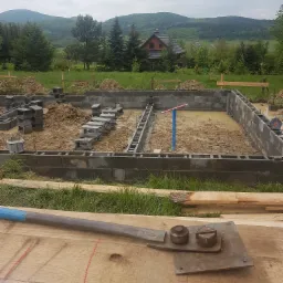 Wykopany fundament z częściowo ułożonymi bloczkami betonowymi, zalany wodą, w tle krajobraz z domem i górami, na pierwszym planie narzędzie do gięcia stali i kalkulator na drewnianym blacie.