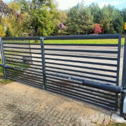 Automatyczna brama przesuwna w kolorze grafitowym, z widocznym siłownikiem i lampą ostrzegawczą, na tle jesiennego krajobrazu z drzewami o zróżnicowanej kolorystyce liści.