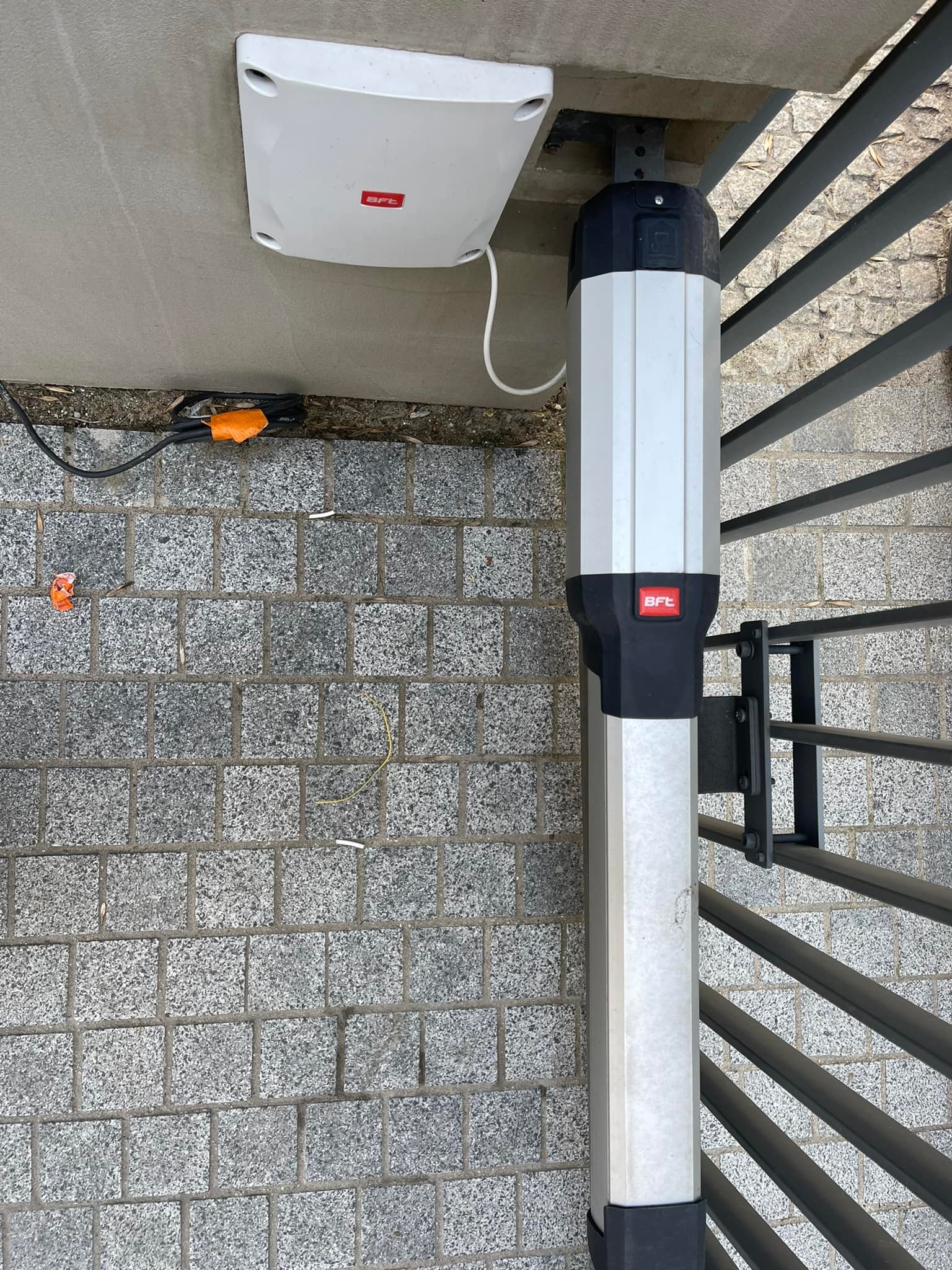 Zewnętrzny napęd do bramy skrzydłowej marki BFT zamontowany na słupku, widoczne okablowanie i kostka brukowa.