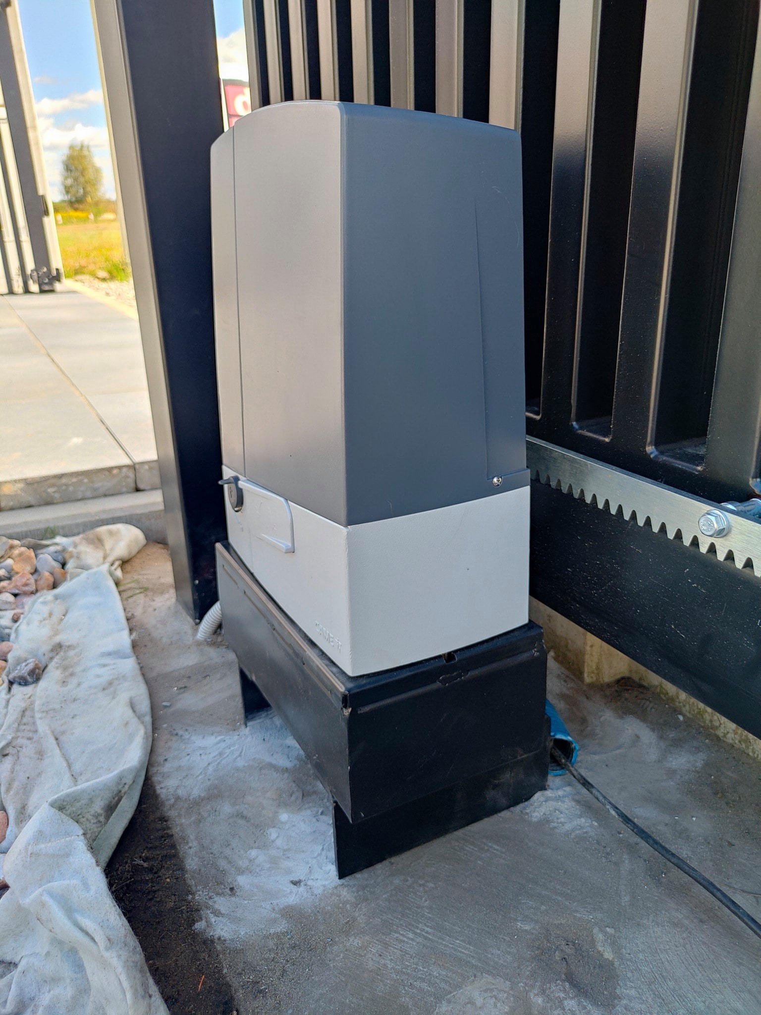 Zamontowany napęd bramy przesuwnej z widoczną listwą zębatą, kabel zasilający w osłonie oraz fragment posesji w tle.