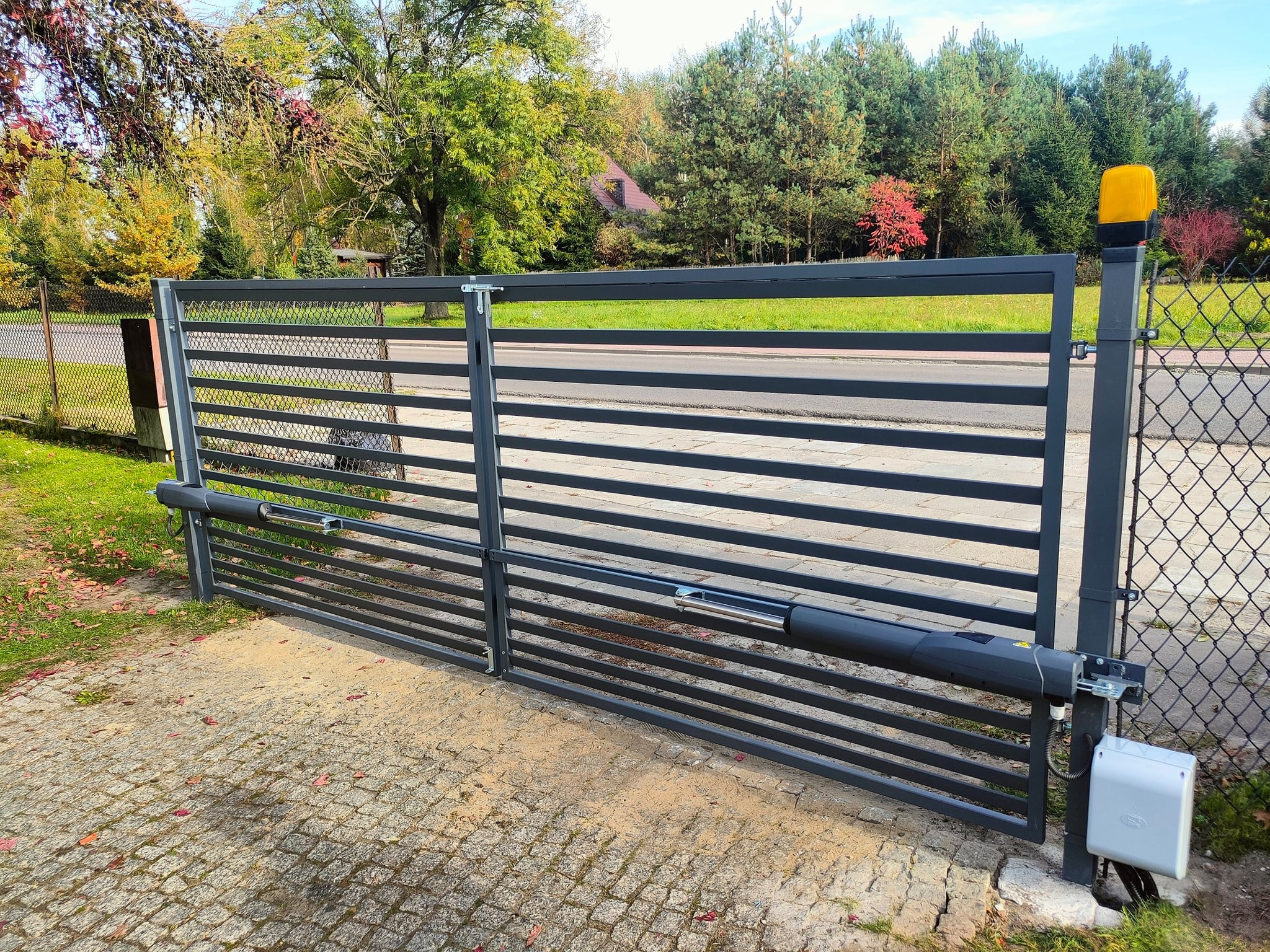 Automatyczna brama przesuwna w kolorze grafitowym, z widocznym siłownikiem i lampą ostrzegawczą, na tle jesiennego krajobrazu z drzewami o zróżnicowanej kolorystyce liści.