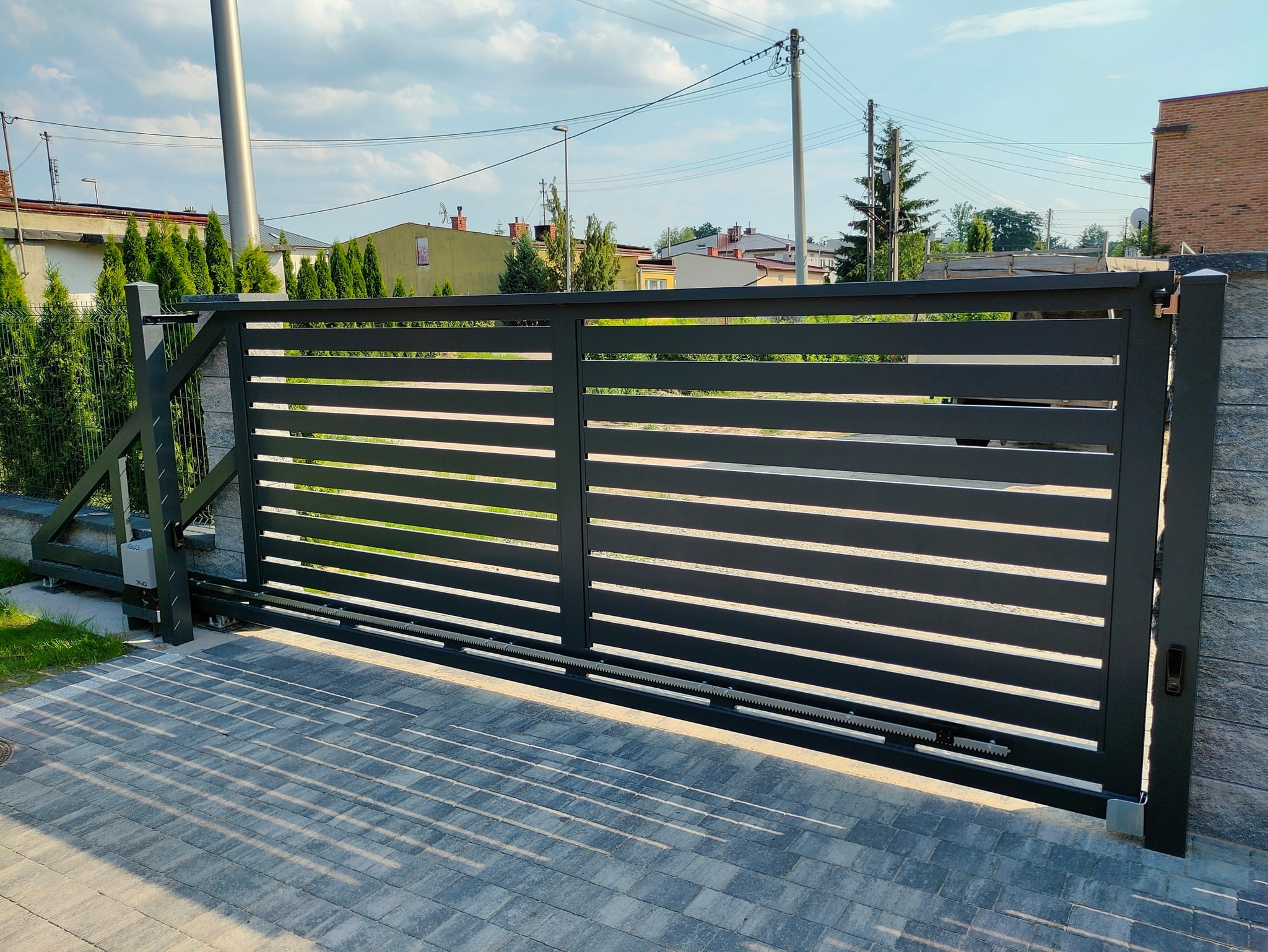Nowoczesna, przesuwna brama w kolorze antracytowym z poziomymi panelami, zamontowana na posesji z kostki brukowej, widoczny napęd i listwa zębata.