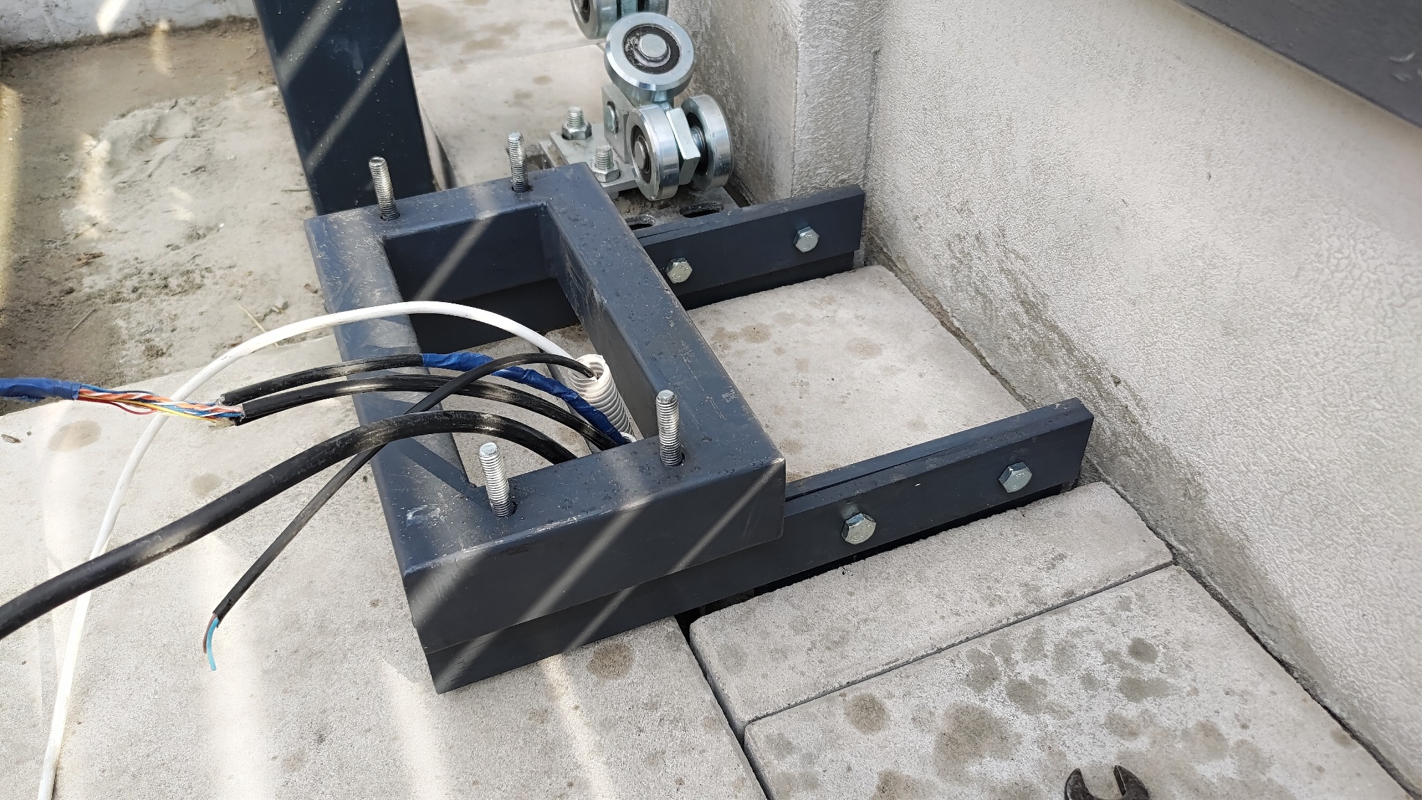Detale montażu automatyki do bramy przesuwnej: czarna metalowa konstrukcja mocująca z widocznymi śrubami, kable elektryczne w trakcie podłączania, betonowa podstawa i fragment ściany.