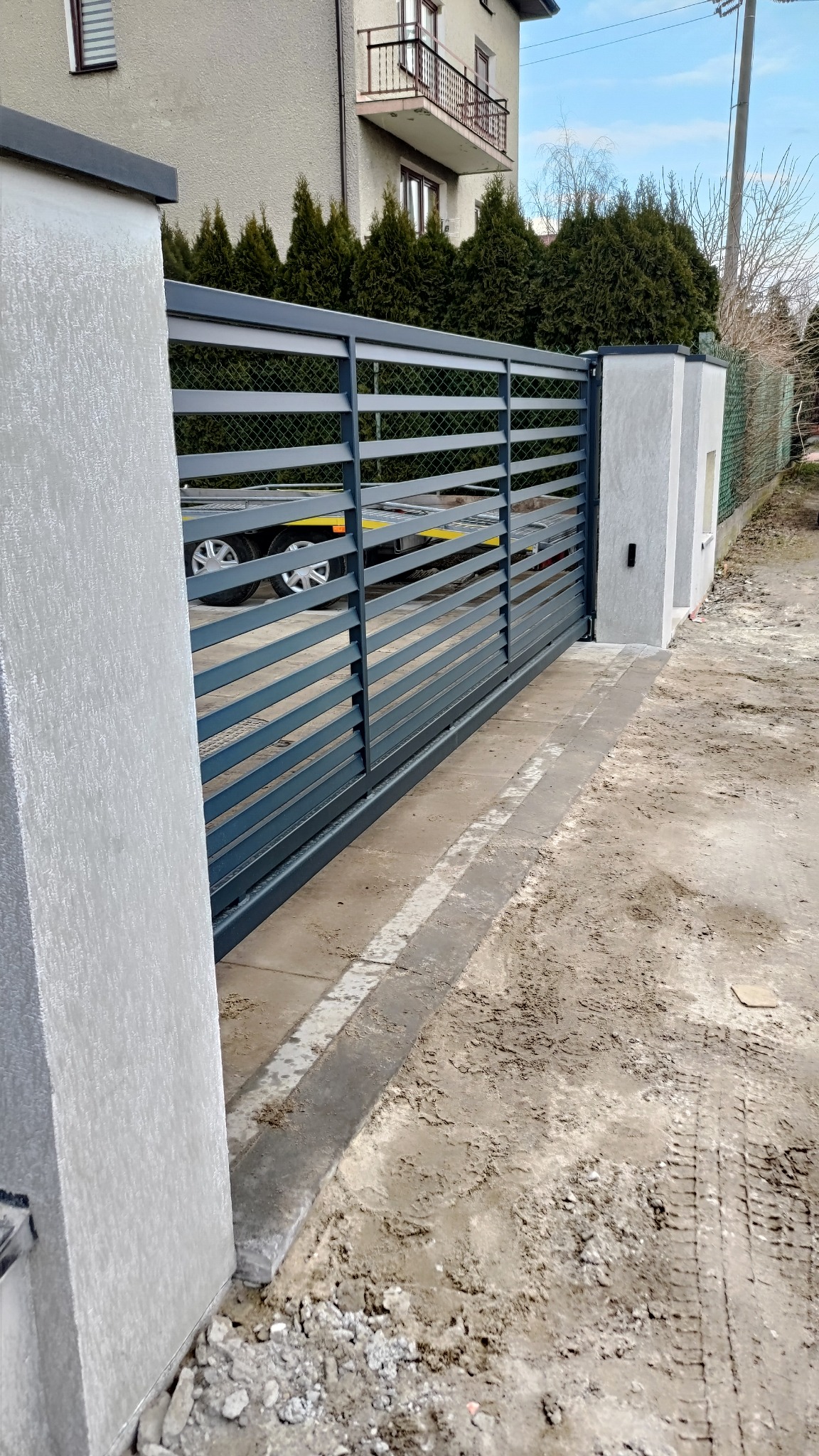 Nowoczesna, przesuwna brama w kolorze grafitowym z poziomymi panelami, zamontowana na betonowych słupkach, widok z zewnątrz posesji, na drugim planie trawnik i budynek mieszkalny.