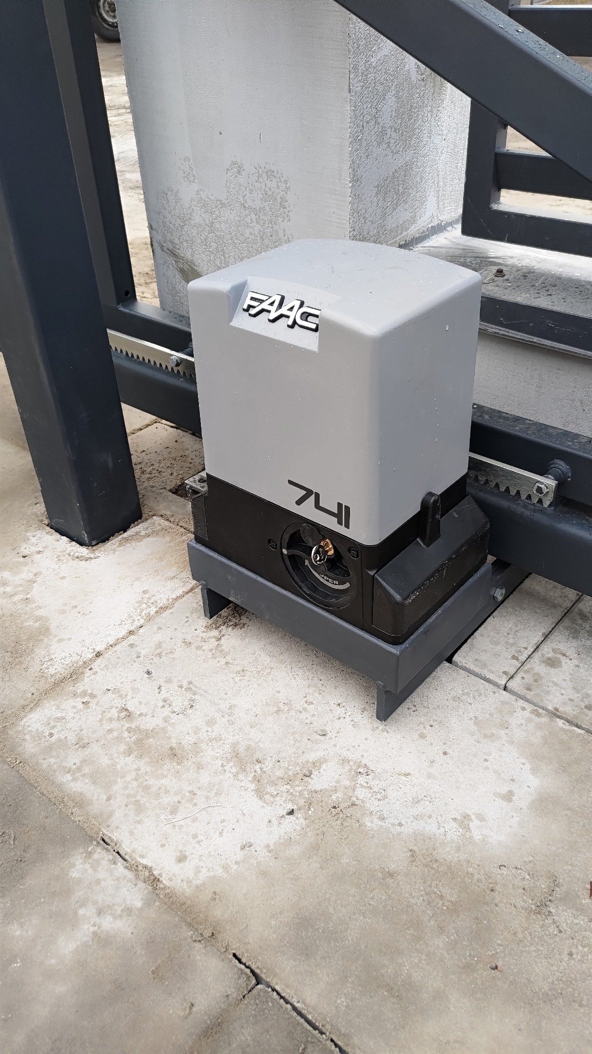 Szary automat do bramy przesuwnej marki FAAC 741 zamontowany na szarej konstrukcji bramy, widoczna zębatka i kluczyk do otwierania awaryjnego.