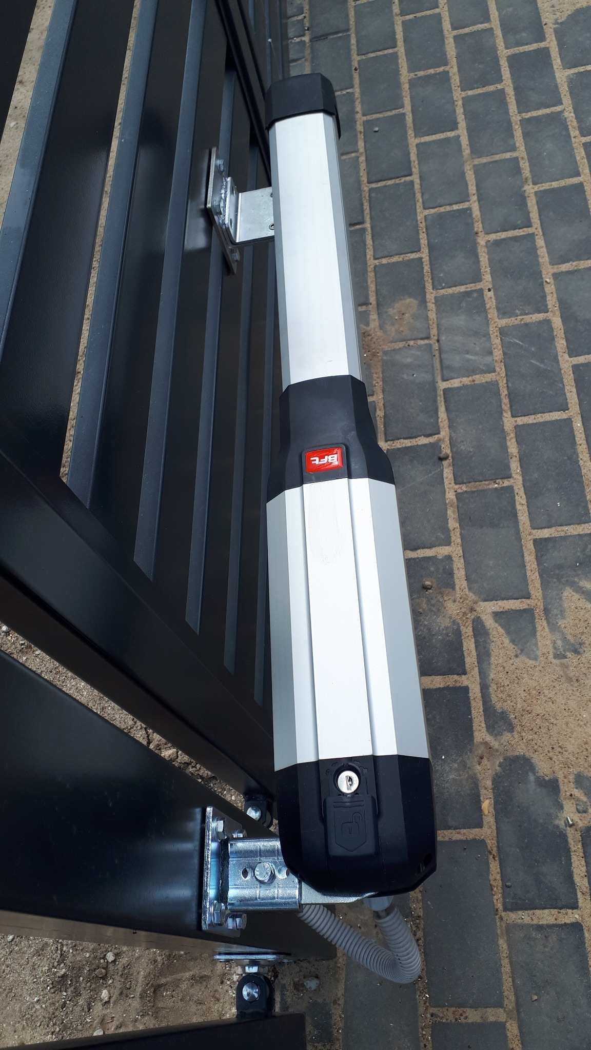 Zamontowany siłownik bramy skrzydłowej BFT z widocznymi mocowaniami, kluczykiem i przewodem zasilającym, na tle kostki brukowej.