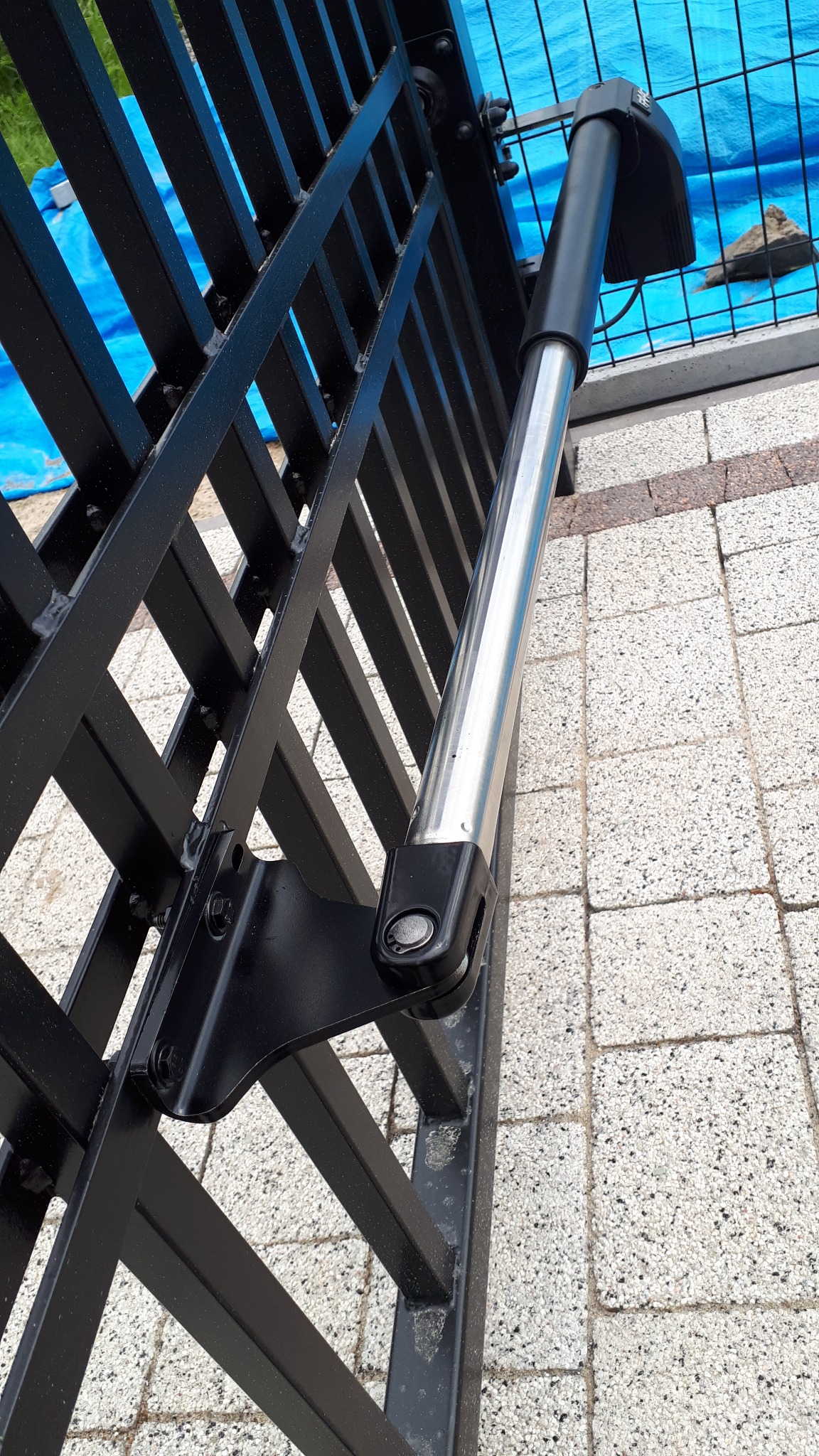 Szczegół mechanizmu automatycznej bramy wjazdowej, widoczny siłownik i czarna metalowa konstrukcja bramy na tle kostki brukowej i niebieskiej plandeki.