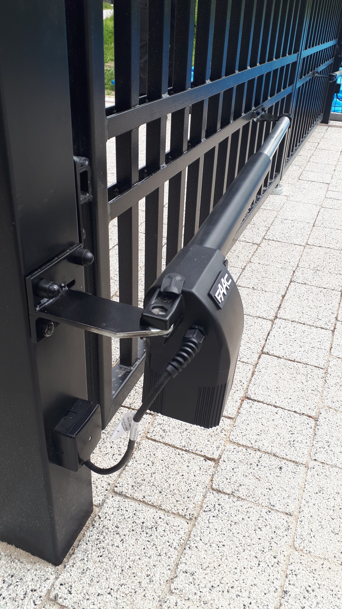 Czarny napęd FAAC zamontowany na metalowej bramie, widoczny kabel zasilający i kostka przyłączeniowa na słupku bramy.