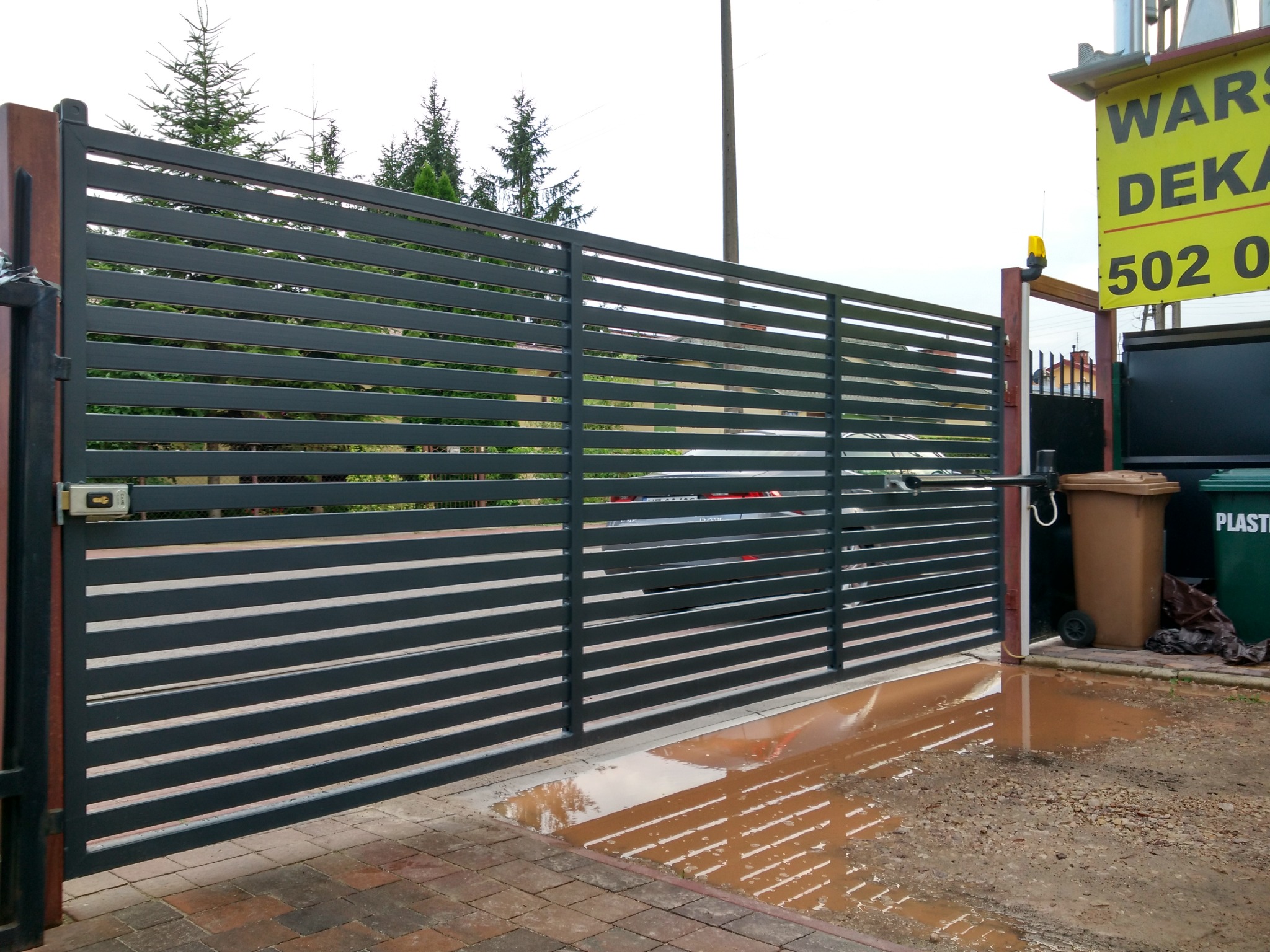 Szara, pozioma brama przesuwna z automatyką, widoczna z zewnątrz posesji, z kałużą na podjeździe i szyldem reklamowym w tle.