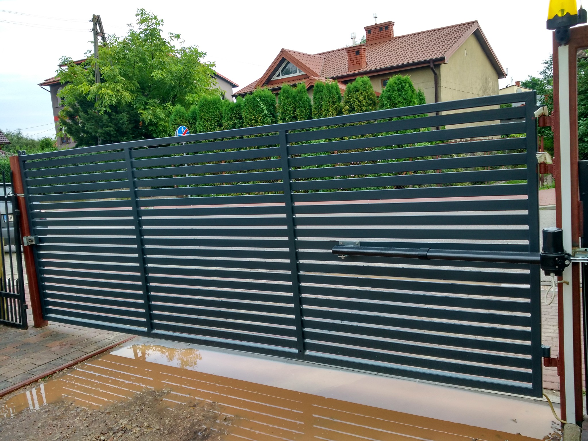 Nowoczesna, przesuwna brama posesyjna z automatyką i poziomymi szczeblami, częściowo otwarta, z widoczną kałużą na podjeździe.