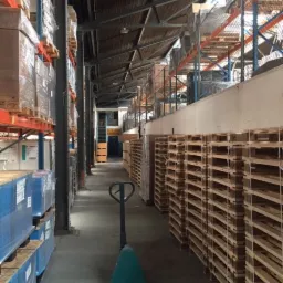 Wnętrze magazynu z regałami wypełnionymi zapakowanymi towarami i stosem palet po prawej stronie, w tle widoczny wózek paletowy.