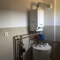 Biały piec gazowy kondensacyjny zamontowany na ścianie, z podłączeniem do zasobnika ciepłej wody, widoczne rury miedziane i elementy instalacji, szaro-biała ściana z gniazdkiem elektrycznym.