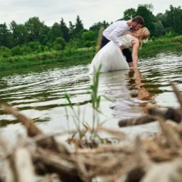Para młoda w wodzie, panna młoda w koronkowej sukni, pan młody w białej koszuli i muszce, pocałunek na tle zieleni i pochmurnego nieba, z bliska widoczne gałęzie na pierwszym planie.