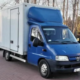 Niebieski samochód dostawczy z białą zabudową kontenerową, zaparkowany na kostce brukowej, widok z przodu na tle drzew bez liści.