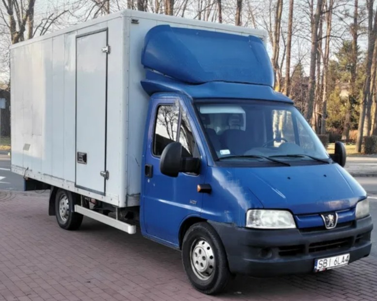 Niebieski samochód dostawczy z białą zabudową kontenerową, zaparkowany na kostce brukowej, widok z przodu na tle drzew bez liści.