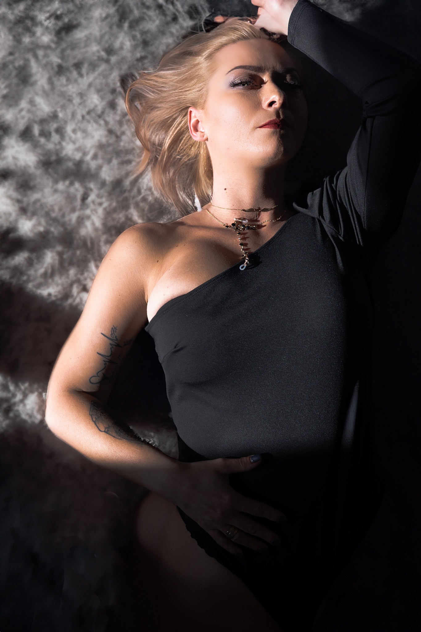 Kobieta w czarnym body leży na futrzanym tle, oświetlona mocnym światłem, z widocznym tatuażem na ramieniu i złotym naszyjnikiem.