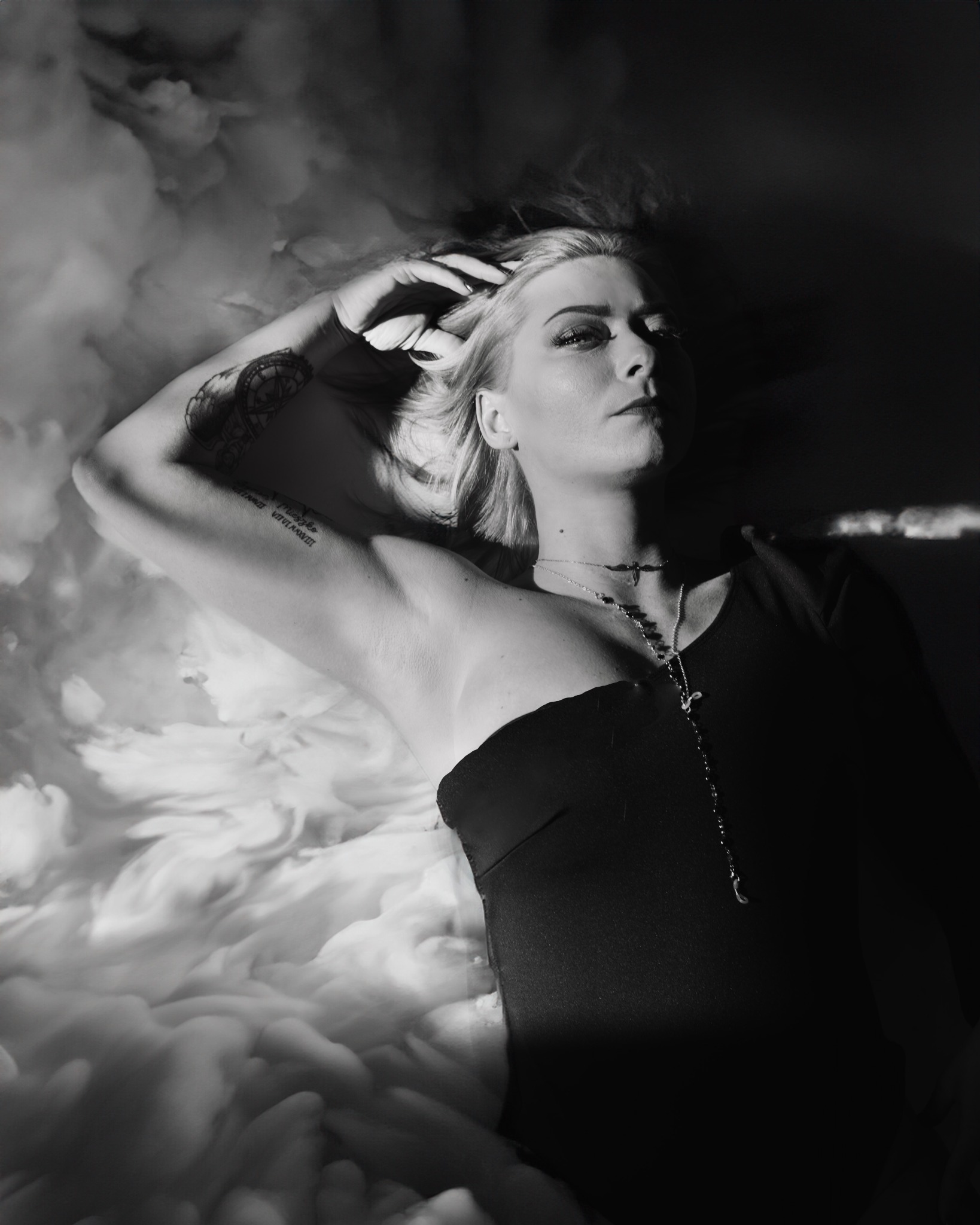 Czarno-białe zdjęcie kobiety leżącej na tle przypominającym chmury, z ręką uniesioną nad głową, widoczne tatuaże na ramieniu i szyi, oświetlona z boku.
