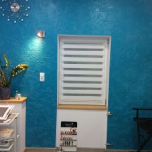 Turkusowa ściana z efektem dekoracyjnym, jasna roleta okienna, fragment blatu z rośliną w doniczce i wózek kosmetyczny w tle. Dekoracyjne wykończenie ściany z widoczną fakturą.