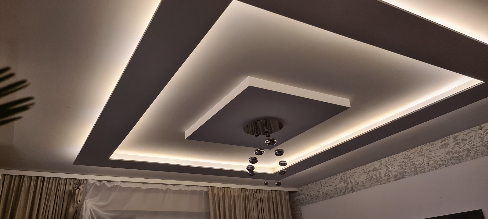 Wielopoziomowy sufit podwieszany z oświetleniem LED i nowoczesnym żyrandolem kulistym, w tle widoczna ściana z dekoracyjną strukturą i zasłony.