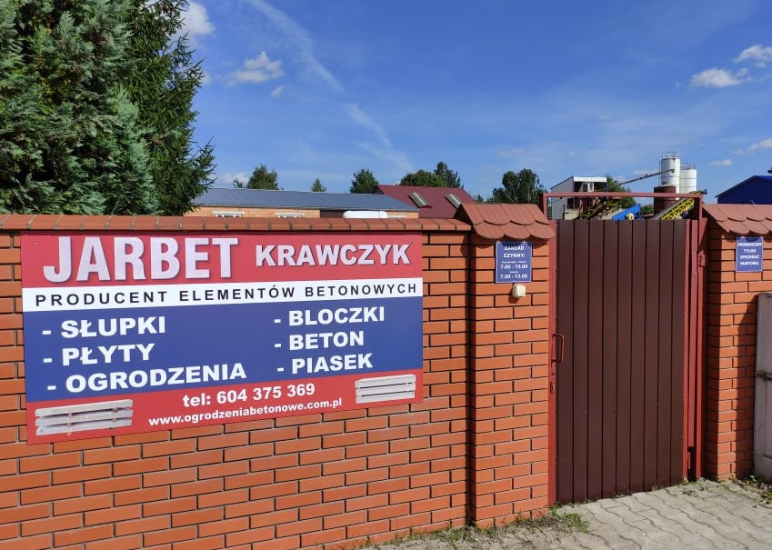 Tablica informacyjna firmy Jarbet Krawczyk, producenta elementów betonowych: słupków, płyt, bloczków, betonu, piasku i ogrodzeń, umieszczona na ceglanym murze wejściowym z brązową bramą.