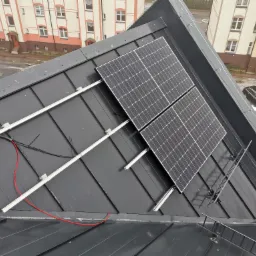 Dwa panele fotowoltaiczne zamontowane na ciemnym, metalowym dachu budynku mieszkalnego, widoczne kable i elementy montażowe.