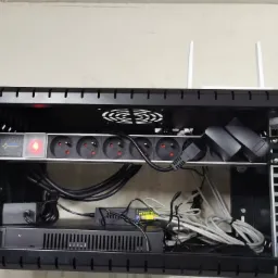 Szafa rack z listwą zasilającą, routerem i okablowaniem, widoczne anteny routera wystające ponad szafę.