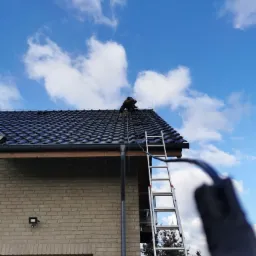 Monter na dachu z ciemną dachówką, instalacja kabli, drabina oparta o ceglany budynek, błękitne niebo z chmurami w tle.